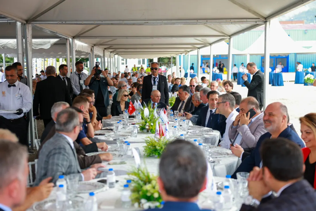 Tören çadırında uzun masalar boyunca oturan davetliler; masalarda Türk bayrakları ve çiçek düzenlemeleri