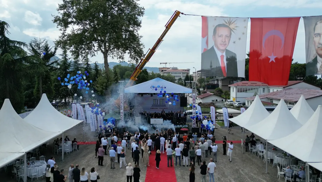 Tören alanı: sahnede konuşma yapılırken havaya konfeti atılıyor, arkada Türk bayrakları ve portreler, önde davetli çadırları
