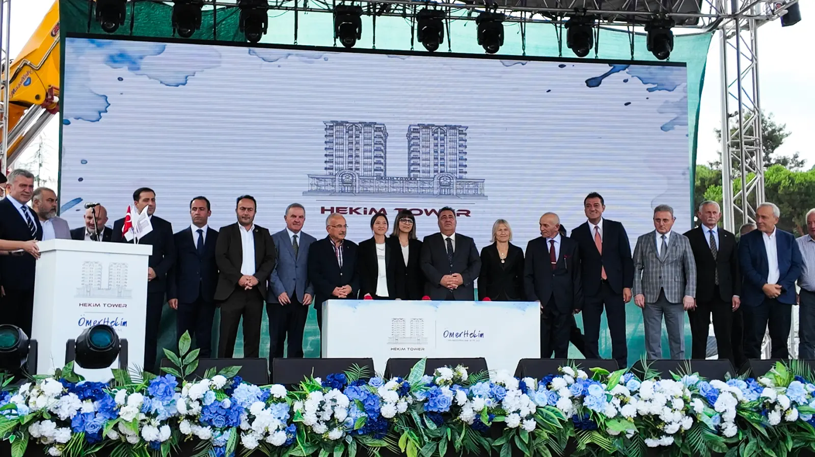 Hekim Tower temel atma töreninde LED ekranda projenin mimari görseli, sahnede protokol sırasında duran davetliler ve önde çiçek düzenlemesi